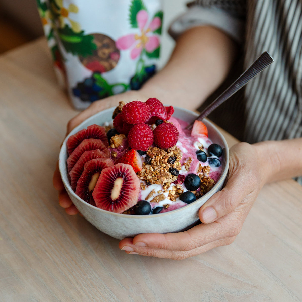 Person holding a bowl of fruit salad with granola, next to a bag of Bountiful Berry & Macadamia Activated Granola.